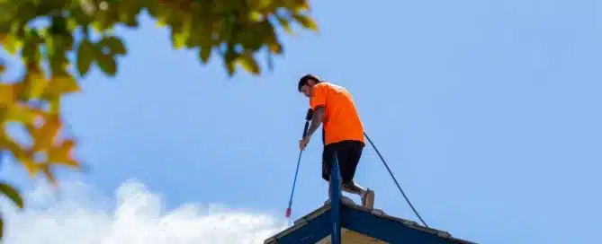 Man power washing a roof. Bright orange shirt, blue trim. "Permacoat roof cleaning.