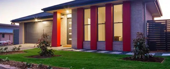 Modern home with brickwork, dark roof, red accents, and manicured lawn. "Ignore Roof Maintenance At Your Own Peril.